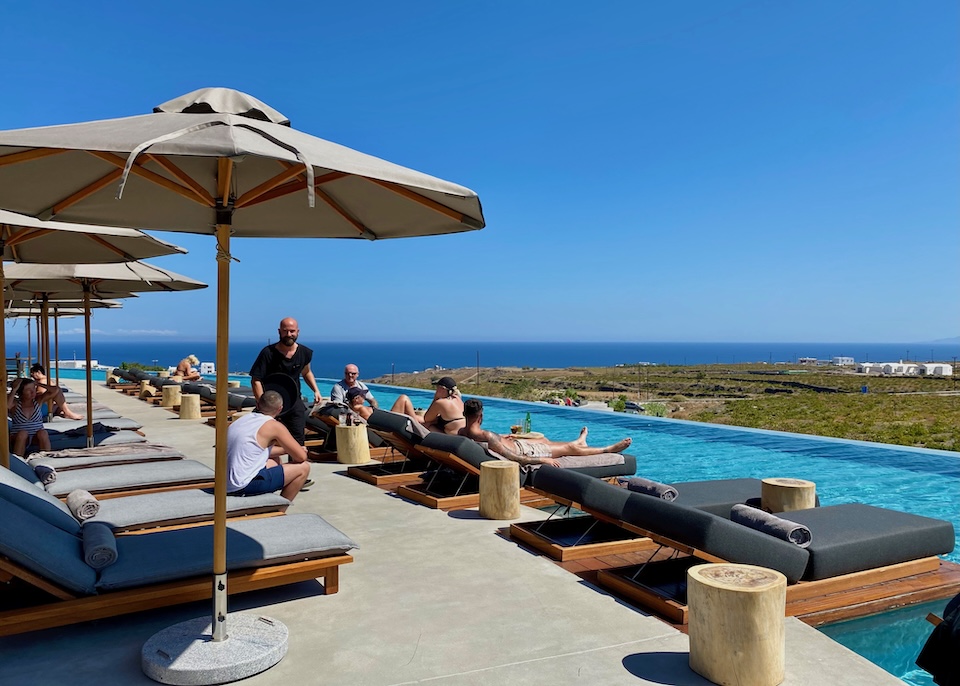 A long infinity pool wraps around a sun deck with sunbeds on the deck and floating on the poolside at Magma Resort in Vourvoulos, Santorini.