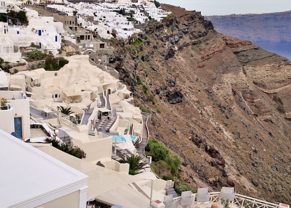 A cream-colored series of cave-style Cycladic buildings centered around a pool with sunbeds on top of the caldera cliffs at Mystique in Oia, Santorini.