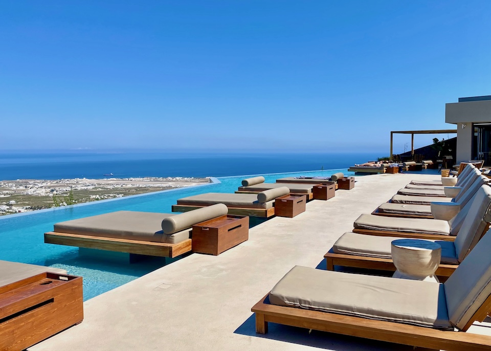 One row of single-sized sunbeds on a terrace and one row of floating, double-sized sunbeds in the infinity pool with a view over the island and sea at North Santorini hotel in Pyrgos village.