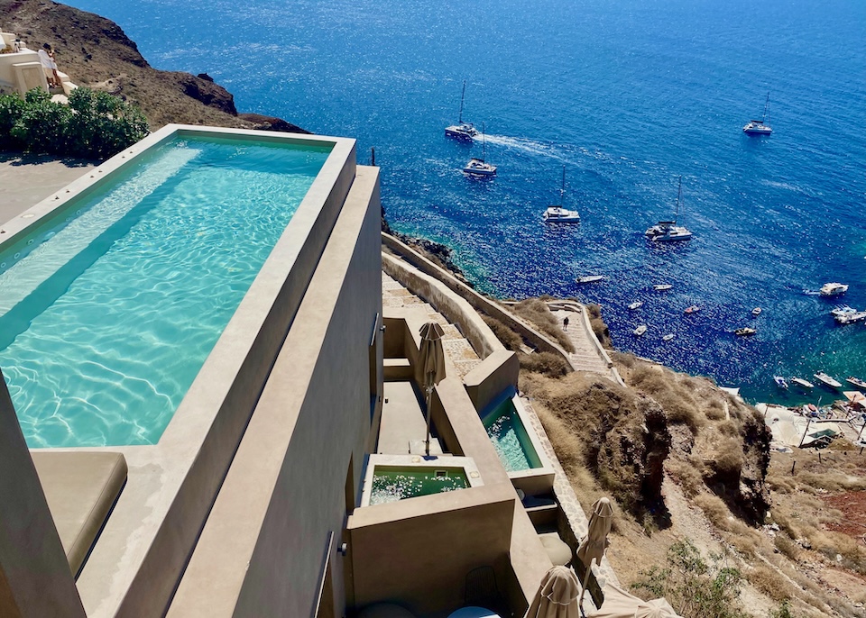 A large pool and two private plunge pool on three terraces down the cliff over the caldera with many boats in the sea at Old Castle Oia hotel in Santorini.