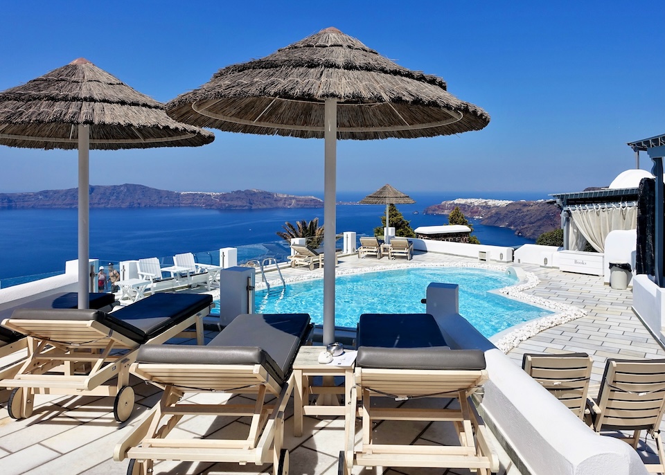 Sunbeds and thatched umbrellas sit behind a pool with views over the caldera toward Oia village and Thirassia Island at Santorini Princess Spa Hotel in Imerovigli.