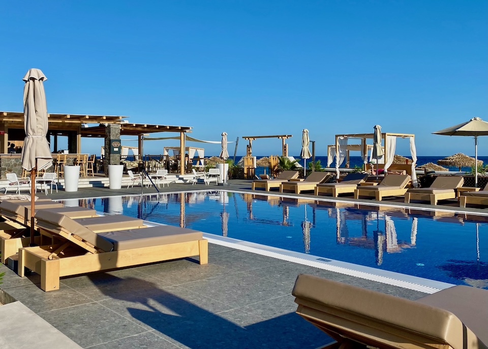 A rectangular pool with sunbeds on its long sides and a bar at the far end with the sea in the background at Sea Breeze hotel in Santorini.
