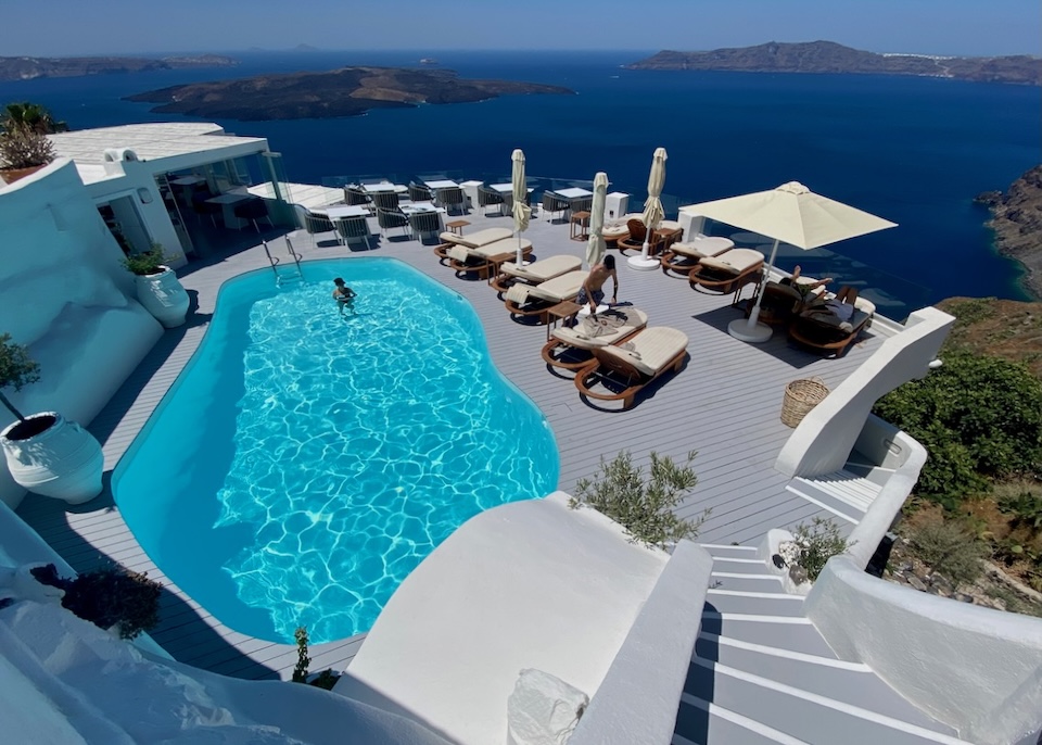 A freeform pool sits at the bottom of a flight of stairs on a wooden deck with sunbeds and a restaurant at Sun Rocks hotel in Firostefani, Santorini.