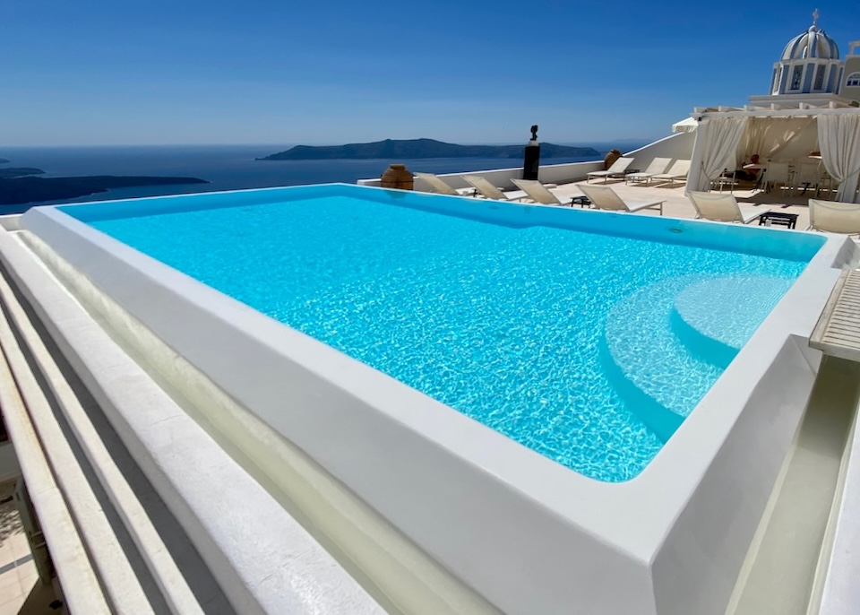 An infinity pool rising above the caldera with a blue and white striped church dome at Tsitouras Collection hotel in Firostefani, Santorini