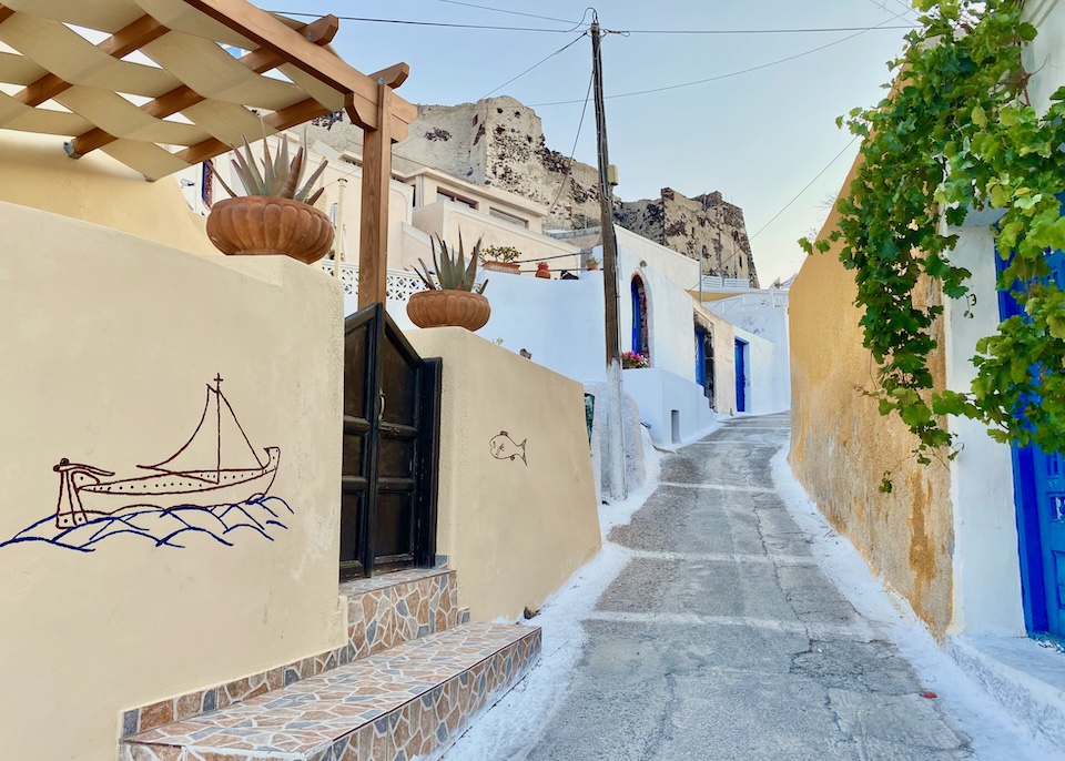 A narrow road leads up to the castle ruins, with yellow and white painted houses on either side, one with a seafaring mural and the other with a blue door and vines in Akrotiri, Santorini.