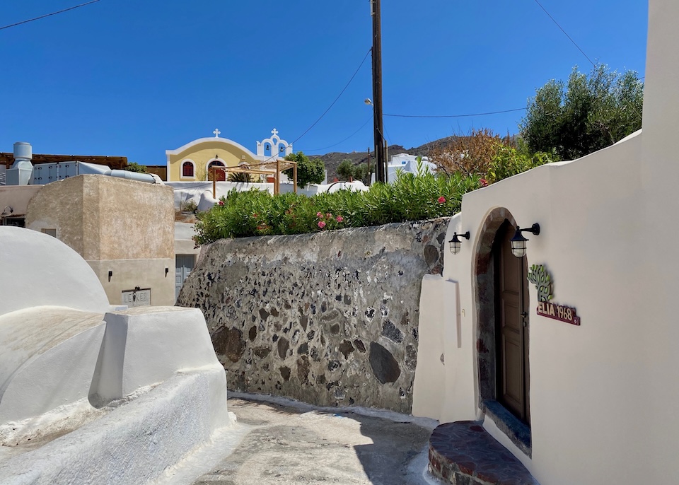 A skinny, curved road leads between low, cave-style homes, hotels, and gardens toward a church in Finikia, Santorini.