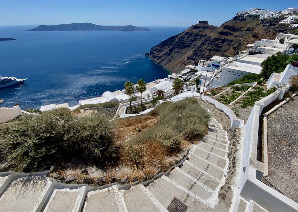 A long, curved stairway leads down through Firostefani village above the caldera with views toward Thirassia Island, Skaros Rock, and Imerovigli village in Santorini.