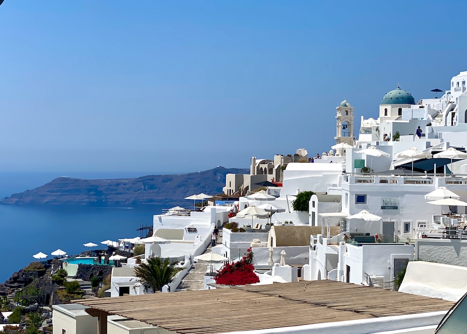 White cubic and barrel-roofed buildings in Imerovigli, Santorini with a blue-domed church and bell tower overlooking the caldera.