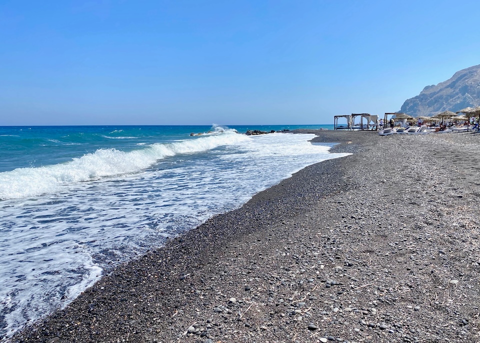 A wide stretch of black sand beach with vibrant turquoise water and a beach club in the background at Kamari Beach in Santorini.