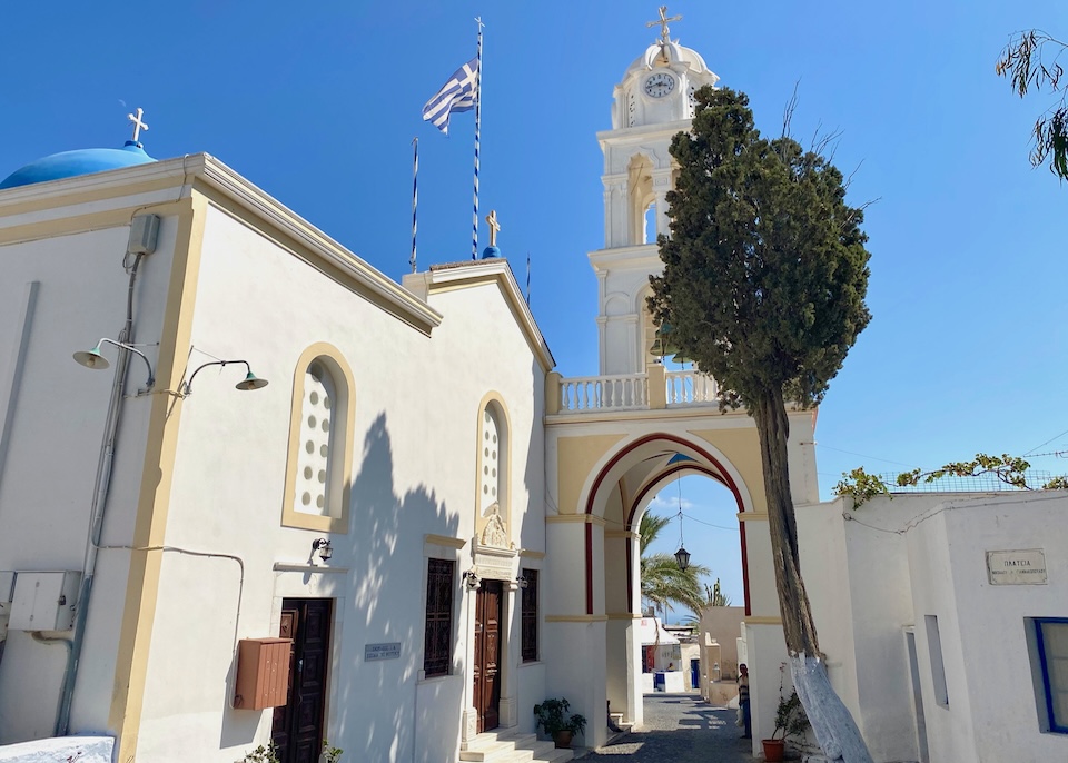 A bell tower with a path carved under it next to a blue-domed church in Megalochori, Santorini.
