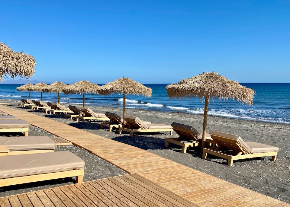 A boardwalk cuts between two rows of sunbeds with thatched umbrellas on a black sand beach in front of a deep blue sea in Perivolos in Santorini.
