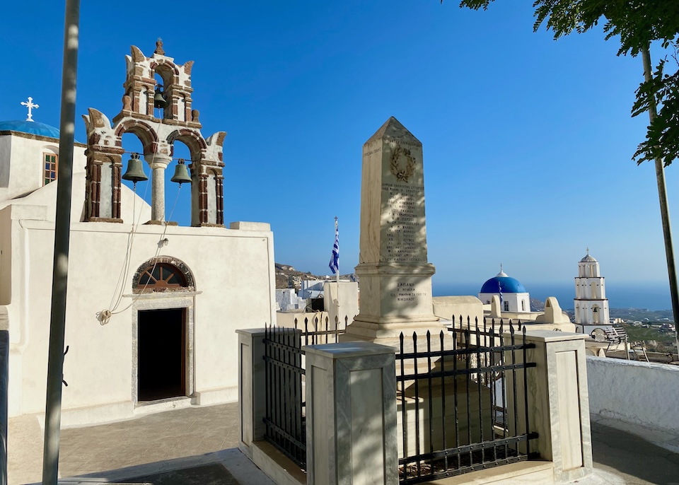 Two blue-domed churches with two bell towers in front of an obelisk-shaped monument sits on the mountainside in Pyrgos, Santorini with the sea spreading below in the distance.