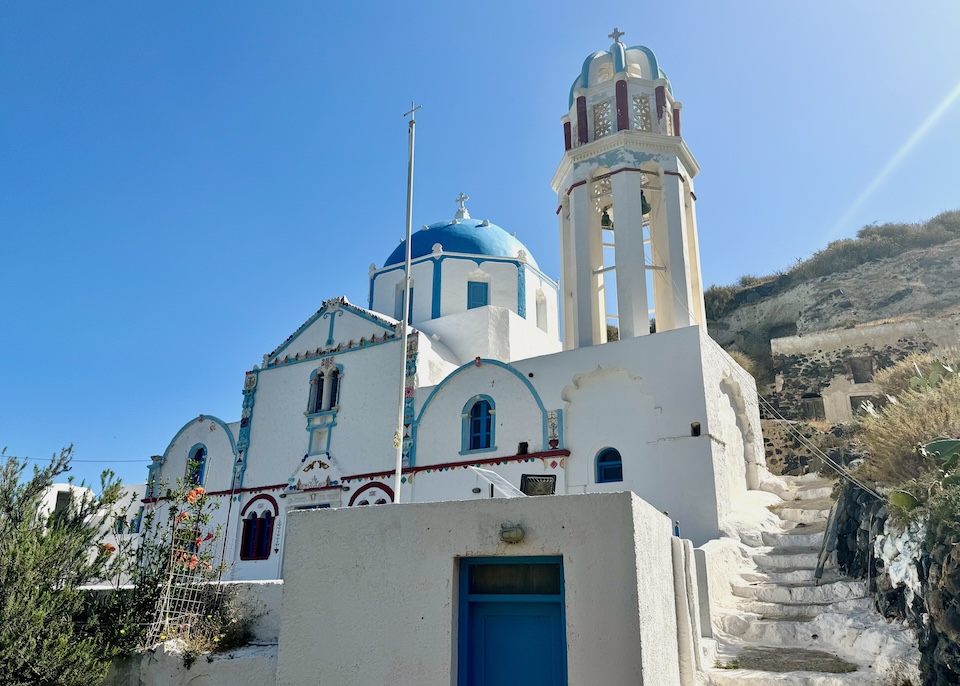An elaborately painted church in red, white, and blue with a dome and bell tower in a deserted village on Thirassia Island across from Santorini