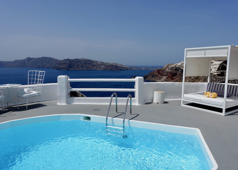 A freeform private pool next to a dining table and Bali bed on a terrace above the caldera at Katikies Kirini hotel in Oia, Santorini.