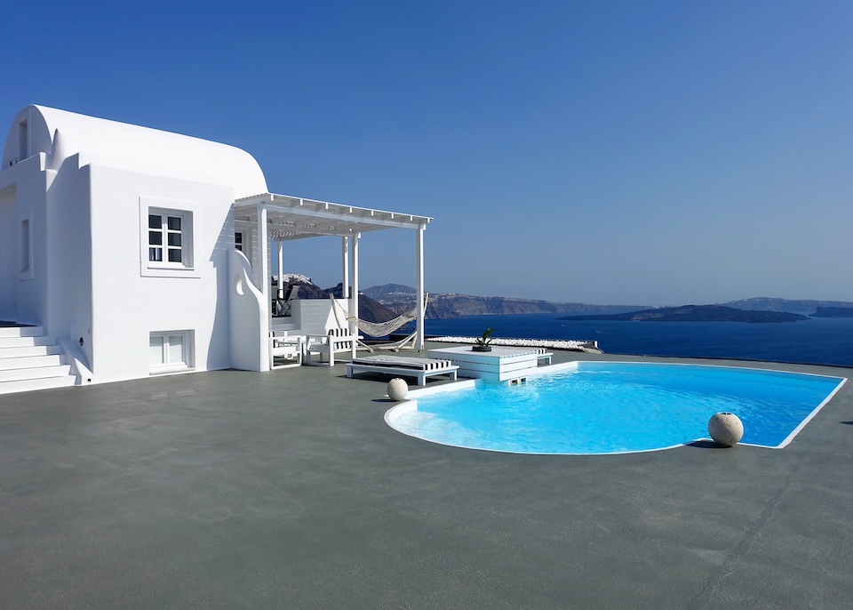 A large private swimming pool on a furnished patio with a pergola and a hammock next to a whitewashed villa overlooking the caldera at Mythique in Oia, Santorini.