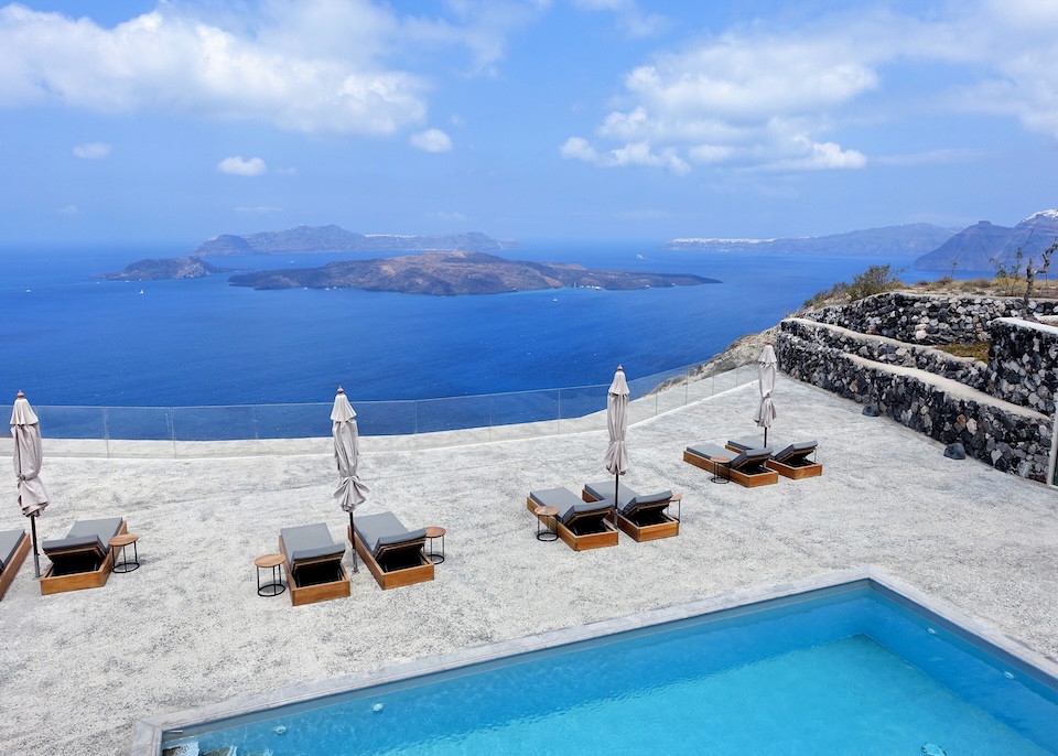 A large private pool on a huge sun deck with four pairs of sunbeds and umbrellas facing the caldera toward the volcanoes at Nafsika Estate in Megalochori, Santorini.