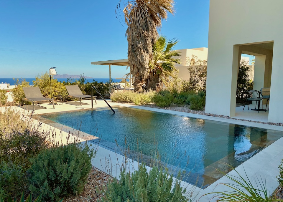 A sunken, infinity edge pool in a garden courtyard with views of the sea in the distance at Nous hotel in Mesaria, Santorini.