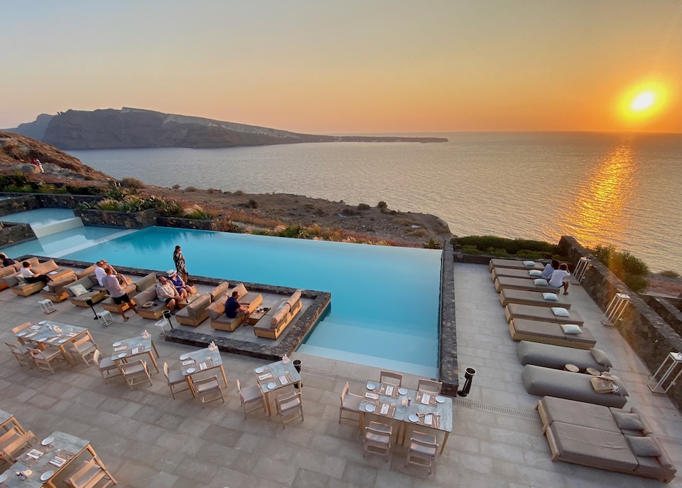 An L-shaped infinity pool with sunbeds on one side and a restaurant on the other overlooks the Aegean Sea at sunset at Canaves Epitome hotel in Oia, Santorini.