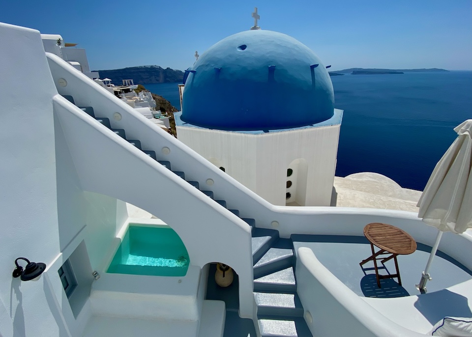 A private plunge pool set under the arch of a staircase immediately behind a blue church dome and the caldera at Oia Spirit Boutique Hotel in Oia, Santorini.