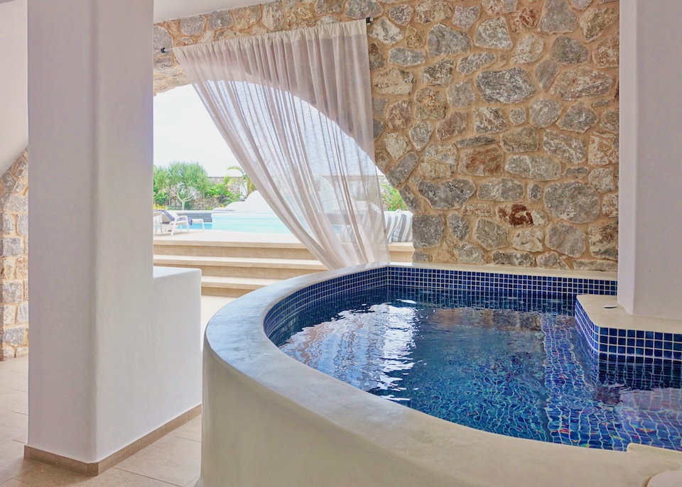 A private jacuzzi in a covered courtyard with a privacy curtain and the main pool in the background at Orabel Suites near Perivolos Beach in Santorini.