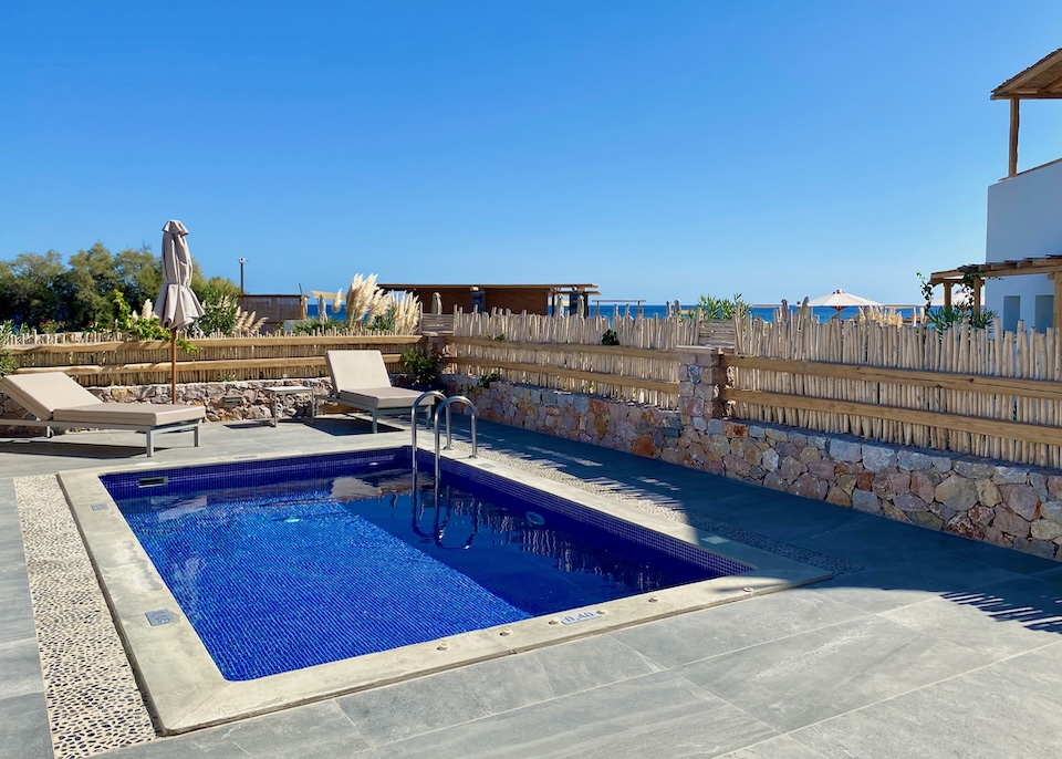 A blue tile private pool on a spacious patio with a pair if sunbeds and a rock and bamboo wall plus the sea in the background at Sea Breeze Beach Resort in Exomitis, Santorini.