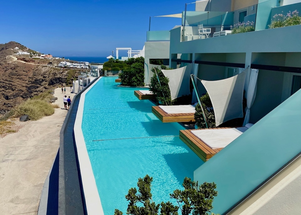 A curved infinity pool divided into sections with ropes and sunbeds hanging over the water at West East Suites in Imerovigli.