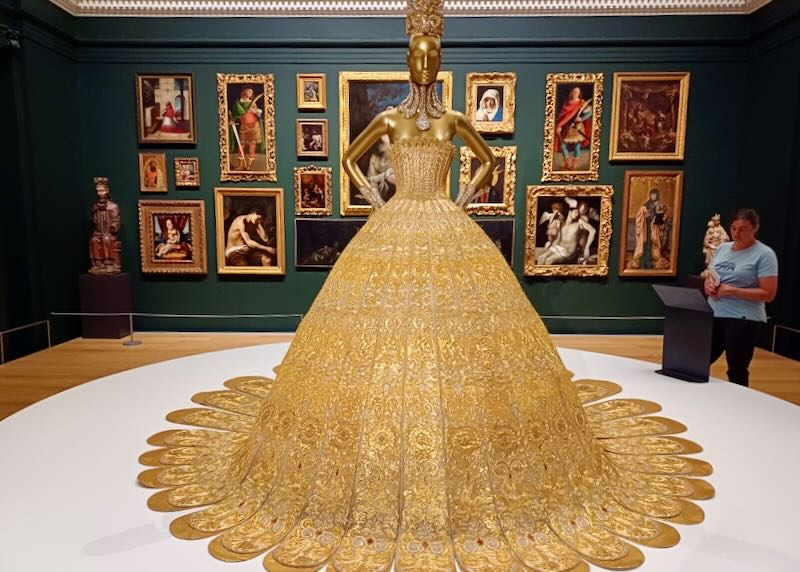 A sculpture of a gold woman in a gold dress, necklace, and crown stands in the middle of a gallery.