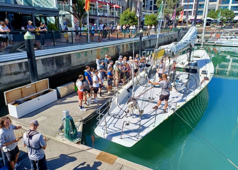 A group of people stand next to a large sailboat in the marina as a few people on board talk to them.