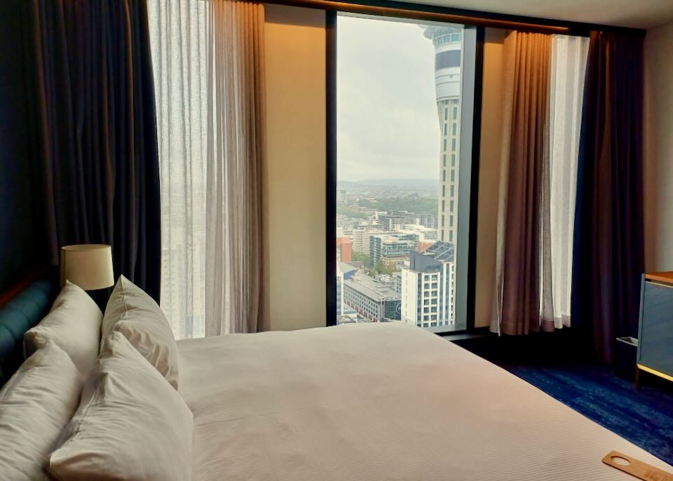 A hotel bed faces a long window showing a tall tower.