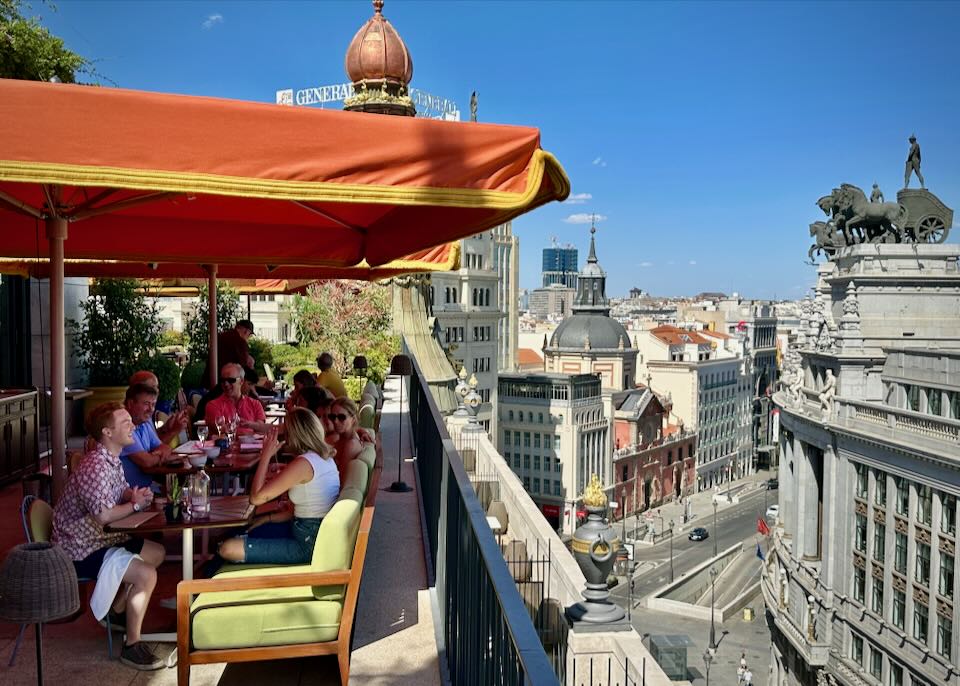 People laugh and dine at an umbrella-covered table on a rooftop terrace overlooking Madrid.