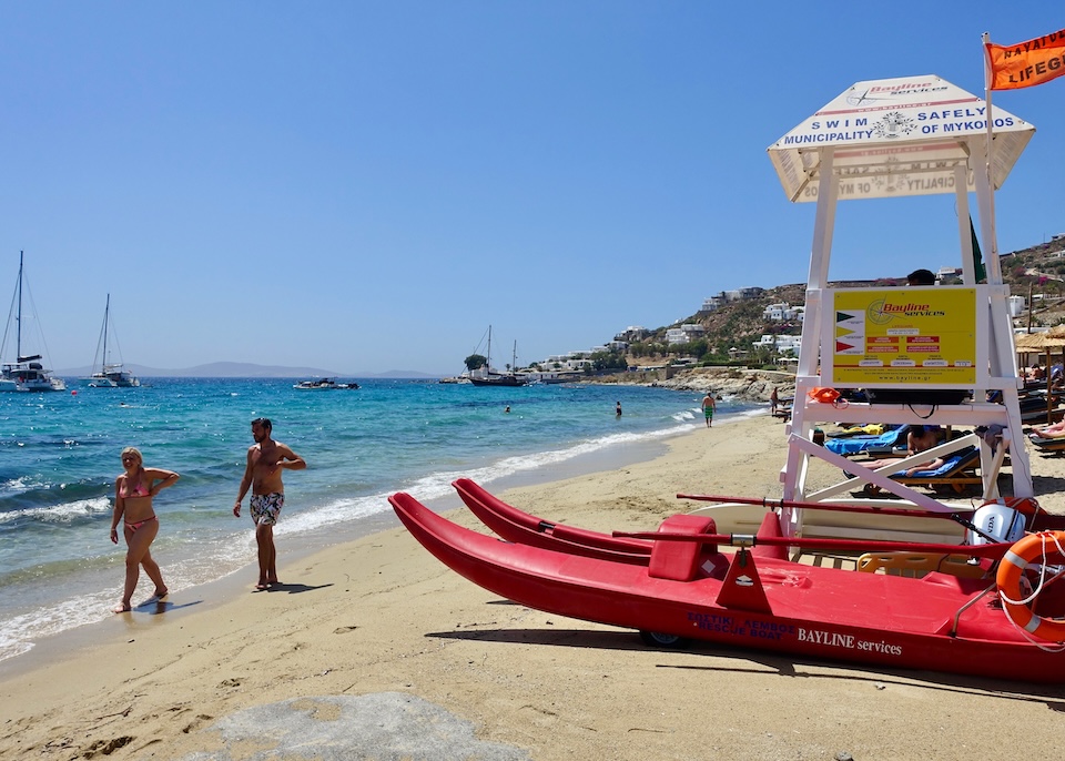 A lifeguard stand next to a red rescue boat on the beach with people strolling on the shore and sailboats in the sea at Agios Ioannis Beach in Mykonos.