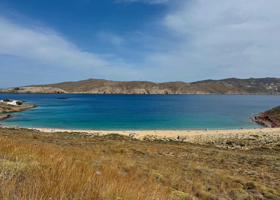 A wide view looking down toward the beach with grasses rolling down the hill at Agios Sostis Beach in Mykonos.