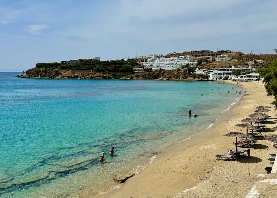 A sandy beach in front of a crescent-shaped bay with a few sunbeds, thatched umbrellas, and people wading into the water at Agios Stefanos Beach in Mykonos.