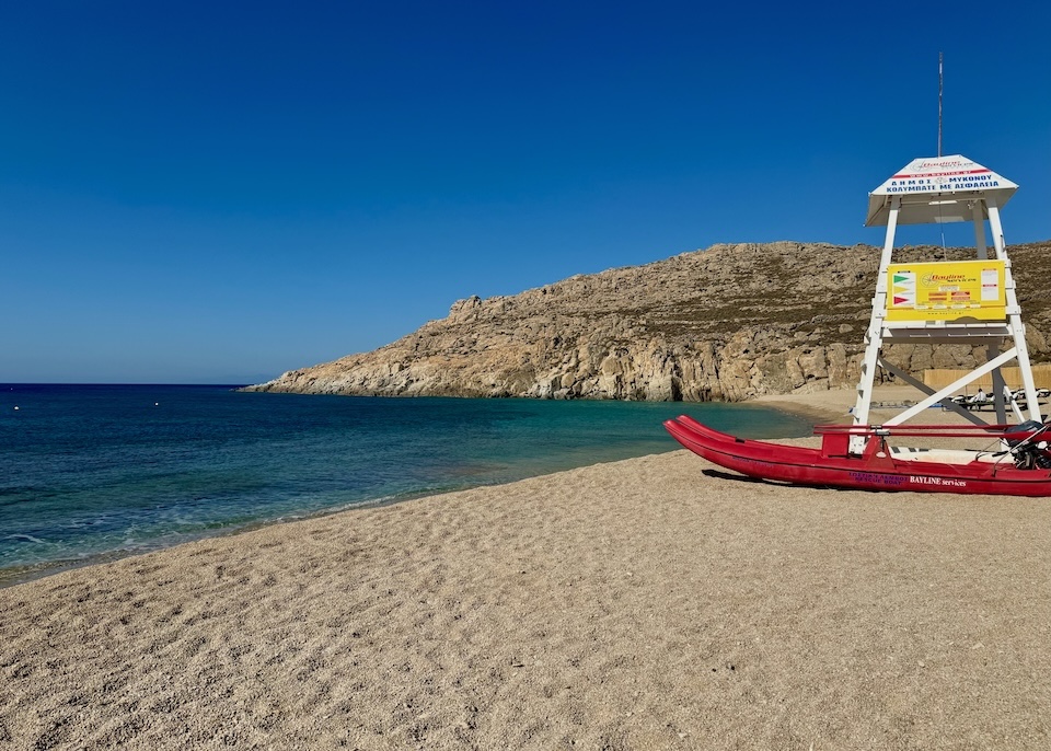 A lifeguard stand and rescue boat sits on a wide, sandy, empty beach with deep blue water at Agrari Beach in Mykonos.