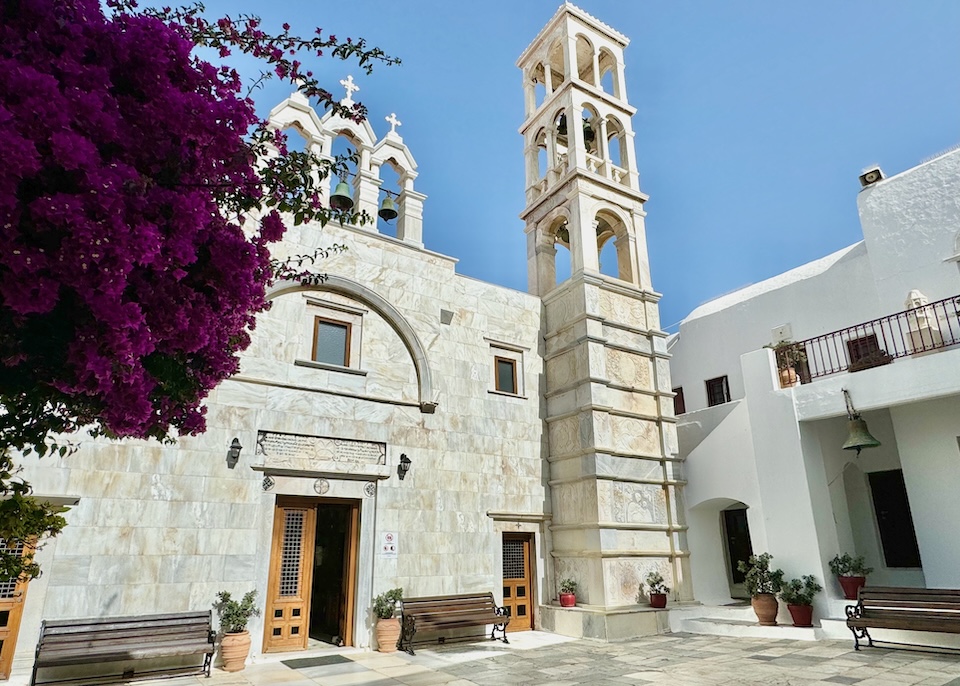 The marble façade of a church and bell tower in a courtyard with a fuschia bougainvillea in front at the church of Panagia Tourliani in Ano Mera village in Mykonos.