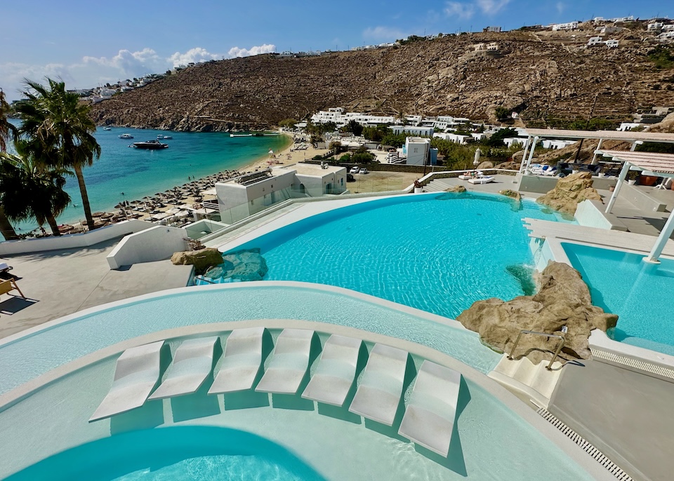 View toward three different shapes and sizes of pool overlooking a beach and sea at Mykonos Blu resort in Psarou.