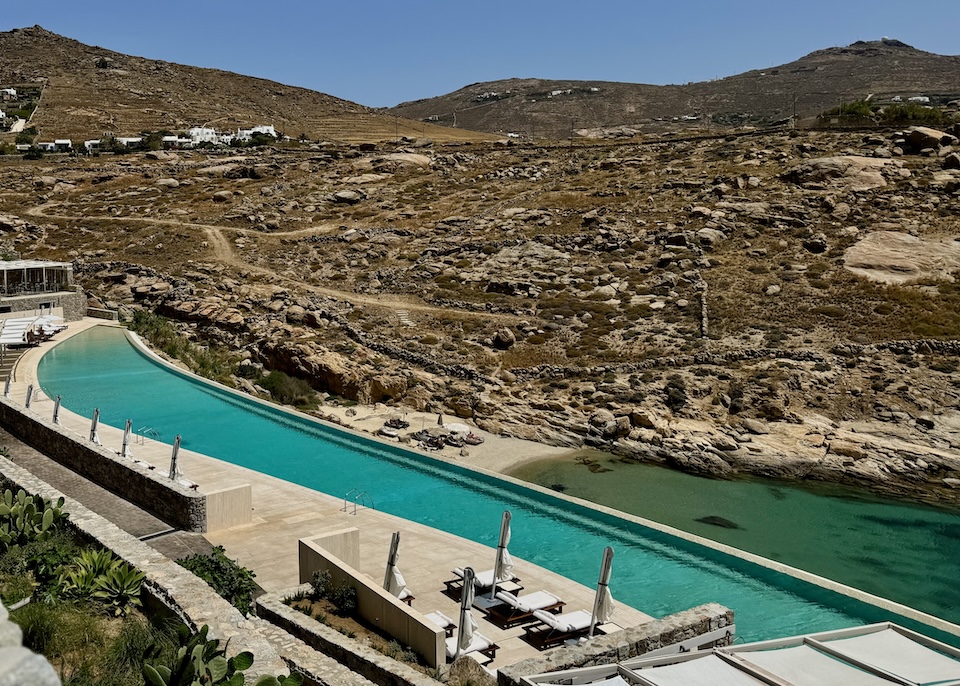 View from above a long, curved pool overlooking a small, private beach with hills rising behind at Cali Mykonos resort in Kalafatis.