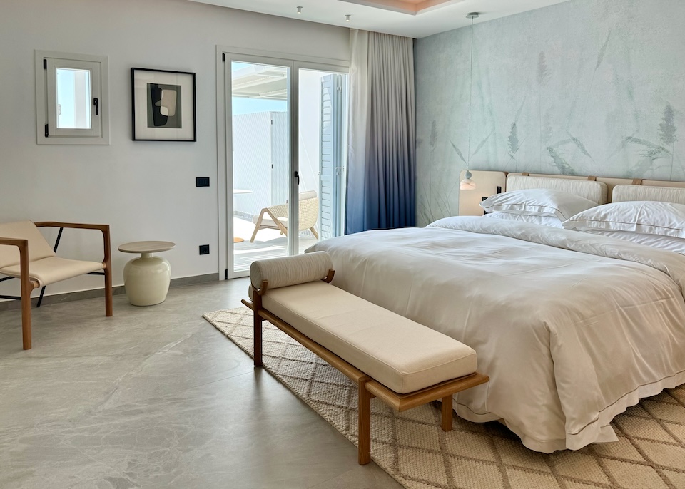 A king-sized bed dressed in white with a nature-ispired wall pattern behind and glass doors leading to a balcony at Deos resort in Mykonos Town.