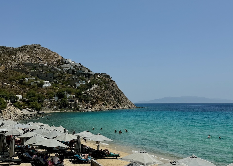 Rows of gray umbrella and sunbed sets on the beach in front of a calm sea with a hill rising behind built up with hotels at Elia Beach in Mykonos.