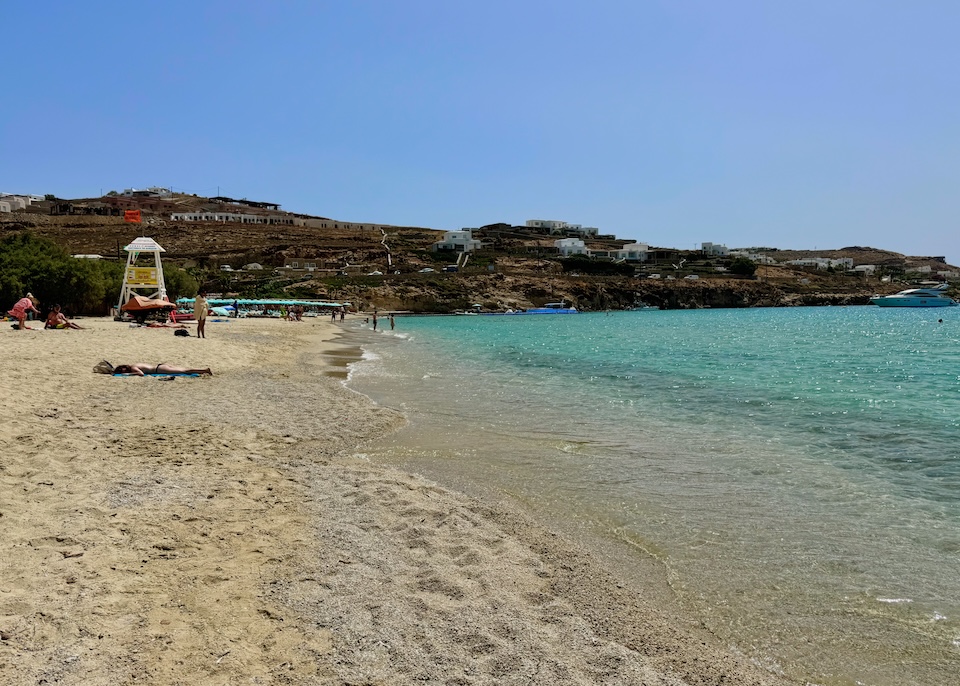 A wide stretch of sand and sea with a beach club and lifeguard stand in the distance and only a few people at Kalo Livadi Beach in Mykonos.