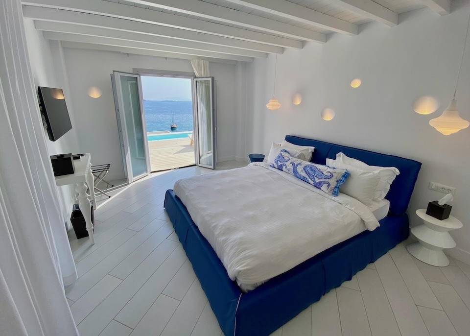 A king-sized bed in white and royal blue in a stark white room with a door leading out to a terrace with a pool and sea view at Katikies resort in Agios Ioannis, Mykonos.