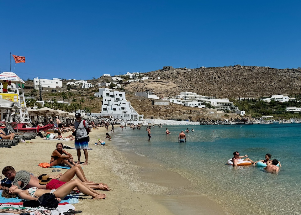 Swimmers and sunbathers enjoy the sand and sea in front of a beach club at Ornos Beach in Mykonos.