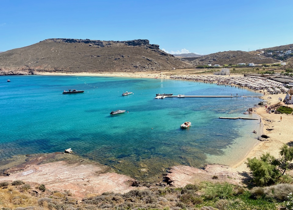 View from a hilltop looking down at Panormos Beach with a busy beach club and a floating pier with a few boats moored in Mykonos.