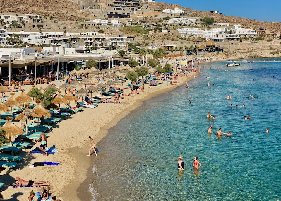 Beach clubs and hotels line the shore and hills, busy with swimmers and sunbathers at Paradise Beach in Mykonos.