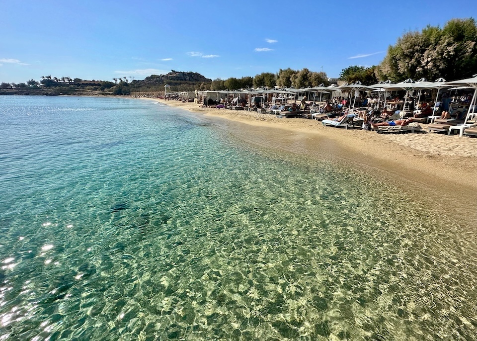 The sea sparkling in the sun in front of a beach club filled with sunbathers at Paraga Beach in Mykonos.