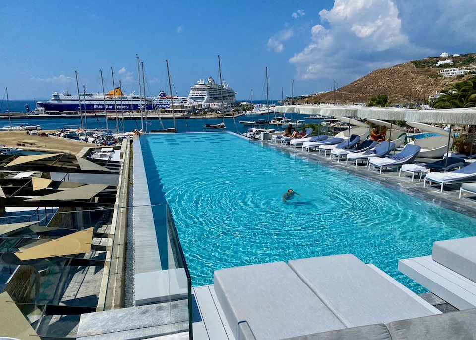 A rectangular infinity pool stretches out toward the sea and ferry port with a row of sunbeds and umbrellas on one side at Mykonos Riviera in Tourlos