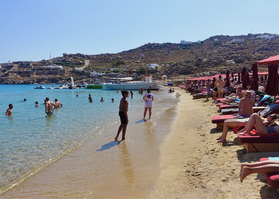 Maroon sunbed and umbrella sets line the shore with swimmers in the sea and a couple of boats landing almost in the sand at Super Paradise Beach in Mykonos.