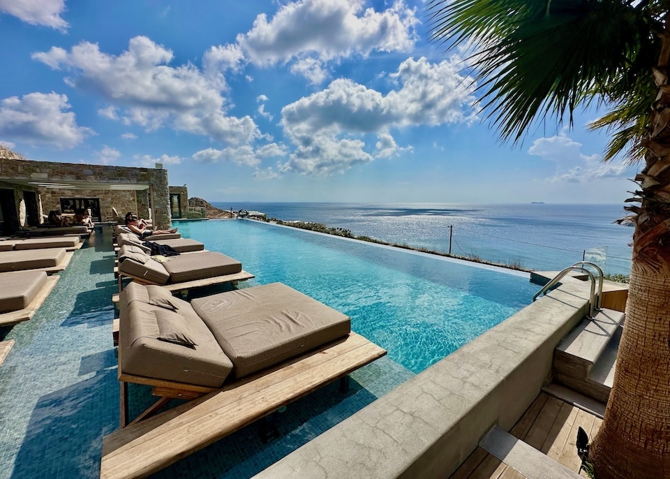 An infinity pool with a row of floating sunbeds inside and a second row of sunbeds on the terrace behind, all facing over the sea at Tropicana Hotel on Paradise Beach, Mykonos.