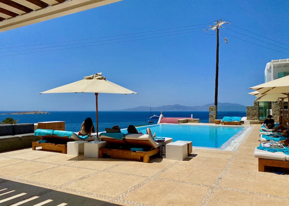 Pairs of sunbeds and umbrellas line two sides of an infinity pool above the sea at Bill and Coo Suites and Lounge in Megali Ammos, Mykonos.