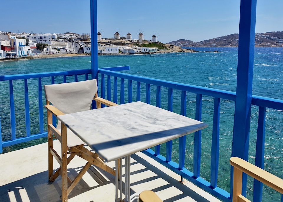 A marble table on a blue painted balcony juts out over the sea with the windmills and Little Venice in view at Bluetopia Suites in Mykonos Town.